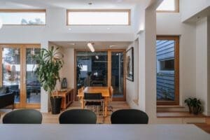 Modern dining area in a home with natural wood finishes, large windows, and contemporary furniture. A wooden dining table with chairs is centered under a pendant light, surrounded by indoor plants, wooden-framed glass doors, and minimal decor. Natural light fills the space through clerestory and side windows.