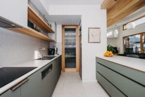 Sleek Australian kitchen by Coys Constructions with sage green cabinetry, open timber shelving, and minimalist tile backsplash.
