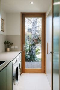 Modern laundry by Coys Constructions with integrated appliances, timber-framed glass door, and garden outlook.