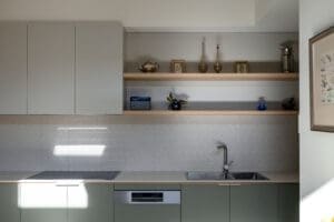 MARIAN HAWTHORN Minimalist kitchen by Coys Constructions with sage cabinetry, open timber shelving, and herringbone tile splashback.